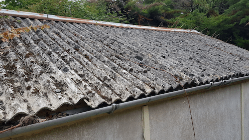 Corrugated cement sheets: these usually contain white asbestos (chrysotile) of which it comprises 10-15% asbestos fibres mixed with cement. There are trees in the backgroun. A metal gutter is at the bottom of the building with a barn wall made from concrete below. The image is taken at an angle. There is a rusted steel trim at the top of the roof.